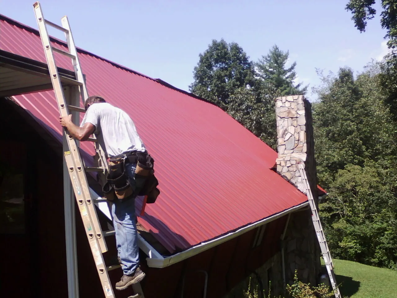 Roofer on a ladder beside a red metal roof and stone chimney — Soffit & Fascia Repair by Roofing Brothers in Cape May Point